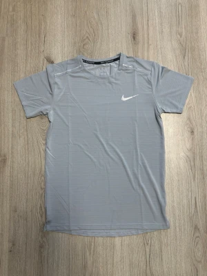 Grå Nike Miller T-Shirt - Säljer en grå kortärmad löpartröja från Nike med diskreta ränder och vit logga på bröstet. Tillverkad i ett lätt och snabbtorkande funktionsmaterial, perfekt för träning och löpning. Rund halsringning och reflexdetalj på axeln.