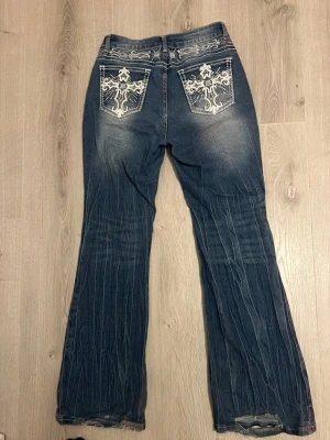 Mörkblå bootcut jeans med brodyr - Säljer ett par mörkblå bootcut jeans med slitningar och vita broderade detaljer på bakfickorna. Jeansen har låg midja, klassisk femficksmodell och kontrastsömmar. Materialet är denim i bomull med lite stretch för skön passform.