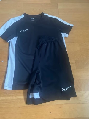 Nike svart Dri-FIT träningsset - Säljer ett svart träningsset från Nike med Dri-FIT-teknologi. Setet består av en t-shirt och shorts, båda med vita detaljer och Nike-logga. Perfekt för fotboll eller annan träning. Materialet är lätt och andas bra.