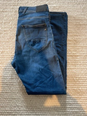 Blå jeans från Replay - Snygga blå jeans från Replay modden annbass med klassisk femficksmodell och diskreta slitningar. Jeansen har normal midja, raka ben och detaljerade sömmar på bakfickan. Tillverkade i mjukt denimtyg som ger en bekväm passform.