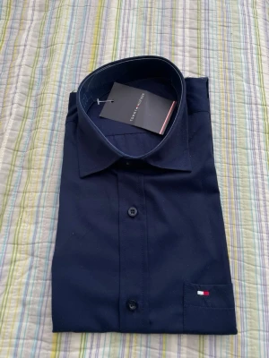 Mörkblå skjorta från Tommy Hilfiger - Stilren mörkblå skjorta från Tommy Hilfiger med klassisk krage och knappar framtill. Liten broderad logga på bröstfickan. Perfekt för dig som gillar en enkel men snygg look.