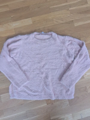 Ljusrosa fluffig stickad tröja - Supermjuk och fluffig stickad tröja i ljusrosa färg. Tröjan har rund halsringning, långa ärmar och ribbade muddar vid hals, ärmslut och nederkant. Perfekt för att hålla sig varm och mysig under kyliga dagar.