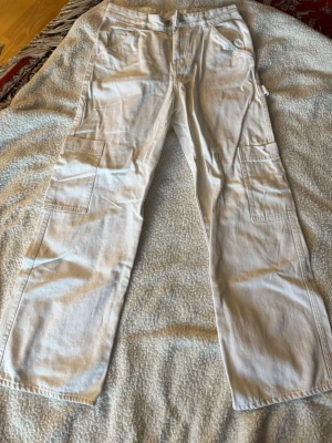 Vita cargobyxor med raka ben - Säljer ett par vita cargobyxor med raka ben och flera praktiska fickor på sidorna. Byxorna har bälteshällor och är tillverkade i ett slitstarkt bomullstyg. Perfekta för en avslappnad och trendig look.