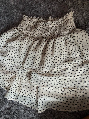 Prickig volangkjol med smock - Supersöt kort kjol med svartprickigt mönster. Kjolen har bred smock i midjan och flera volanger, den är vit/beige aktig och är i storlek XXS