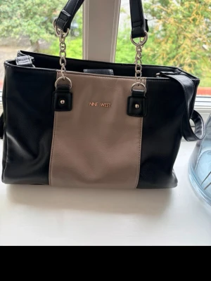 Svart och beige handväska Nine West - Snygg handväska från Nine West i svart och beige konstläder. Väskan har silverfärgade kedjedetaljer, dubbla handtag och en justerbar axelrem. Rektangulär form med gott om plats för dina grejer. Perfekt accessoar för en trendig look.