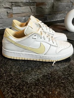 Nike Dunk Low vit/beige sneakers - Nike Dunk Low sneakers i vitt och beige med klassisk Swoosh på sidan. Skorna har lågt skaft, perforerad tåbox och beige detaljer på häl och sula. Tillverkade i läder med textilfoder och snörning framtill. Perfekta för en clean och stilren look.