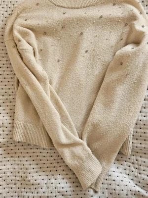 Beige stickad tröja med pärlor - Mysig beige stickad tröja med dekorativa pärlor över hela framsidan. Tröjan har rund halsringning, långa ärmar och ribbade muddar vid ärmslut och nederkant. Dock nopprig pga användning