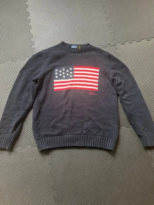 Mörkblå stickad tröja med USA-flagga - Mörkblå stickad tröja med en stor amerikansk flagga i rött, vitt och blått på bröstet. Tröjan har rund hals, ribbade muddar och RL-broderi under flaggan. Perfekt för dig som gillar klassisk och stilren design.