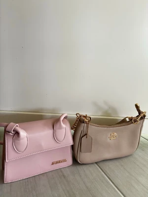 Två trendiga handväskor i pastell - Säljer två snygga handväskor: en ljusrosa Jacquemus med unik miniform och topphandtag, samt en beige väska med gulddetaljer och kedjerem. Båda är perfekta accessoarer för att lyfta din outfit och har en modern, stilren design. Skriv vilken som intresserar dig! Kan diskutera pris!