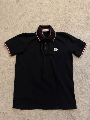 Svart Moncler pikétröja - Klassisk svart pikétröja från Moncler med korta ärmar och krage med röda och vita ränder. Tröjan har knappslå framtill och Moncler-logga på bröstet. Perfekt till jeans eller shorts för en clean och stilren look.