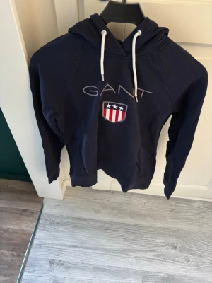 Mörkblå hoodie från GANT - Snygg mörkblå hoodie från GANT med vit dragsko i huvan och broderad logga samt sköld med stjärnor och ränder på bröstet. Klassisk passform och mjukt material som känns skönt mot huden. Perfekt för en avslappnad och stilren look.
