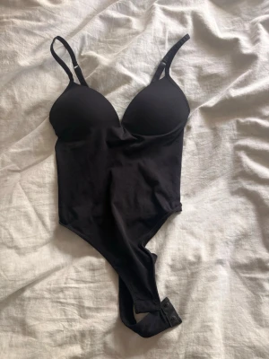 Svart body från gina  - Snygg svart body med justerbara axelband och vadderade kupor för extra stöd. Bodyn har tryckknappar i grenen för enkel av- och påtagning. Tillverkad i mjukt stretchigt material som sitter skönt mot kroppen.
