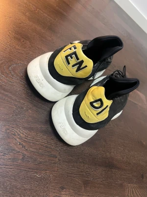 Svarta Fendi sneakers med gul detalj - Unika svarta sneakers från Fendi med chunky vit sula och gul detalj på hälen där det står 'FENDI'. Skorna har en modern design med elastisk snörning och är tillverkade i mocka och textil. Perfekta för dig som vill sticka ut med en lyxig streetstyle.