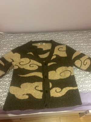 Grön kofta med beige molnmönster - Unik grön kofta med beige molnliknande mönster och v-ringad hals. Koftan har knäppning framtill och är gjord i ett fluffigt, stickat material som känns riktigt mysigt. Koftan är från woodbird