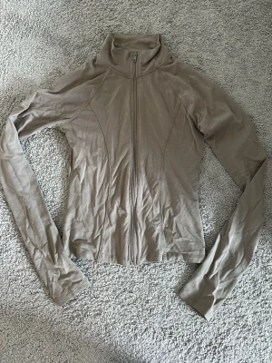 Beige zip träningströja - Slimmad beige ziptröja med hög krage och långa ärmar. Tröjan har en hel dragkedja framtill och är tillverkad i ett tunt, stretchigt material som sitter tajt mot kroppen. Perfekt att dra över en träningstopp. 