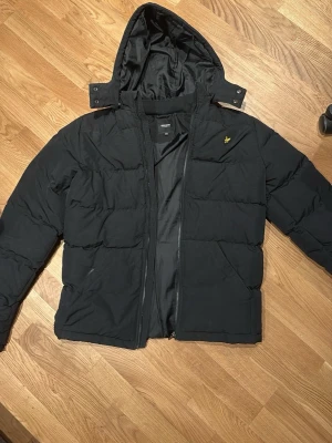 Svart pufferjacka med huva - Säljer en svart pufferjacka från Lyle & Scott med avtagbar huva och dragkedja framtill. Jackan har ett guldfärgat logomärke på bröstet och två sidofickor. Perfekt för kalla vinterdagar och riktigt skön att bära.