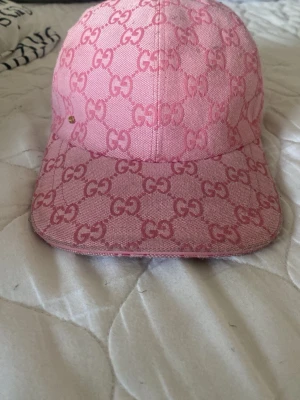 Rosa keps med GG-mönster från Gucci - Säljer en rosa keps från Gucci med ikoniskt GG-mönster över hela kepsen. Kepsen har böjd skärm och justerbart spänne bak i matchande rosa. Materialet är canvas med tydlig struktur och kepsen har en lyxig vibe.