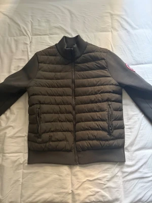 Mörkgrön canada goose cardigand - Snygg mörkbrun dunjacka med quiltad front och ribbade muddar. Jackan har hög krage, två fickor med dragkedja och långärmade stickade ärmar. Perfekt för kyliga dagar och har en stilren look med diskret logga på ärmen.