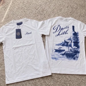 Två Vita t-shirt med blått tryck - Vit t-shirt med blått tryck på ryggen som föreställer en vinflaska, glas och naturmotiv samt texten 'Dessas Lith'. Framtill står det 'Marcel' i blått. Klassisk passform och rund hals.