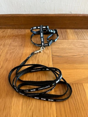 Sele till kanin  - Svart sele och koppel med kaniner på. Koppel: 127 cm Sele: Storlek: XS/S Ryggband: 6,5 cm Halsband: 18-25 cm Magband: 24-35 cm