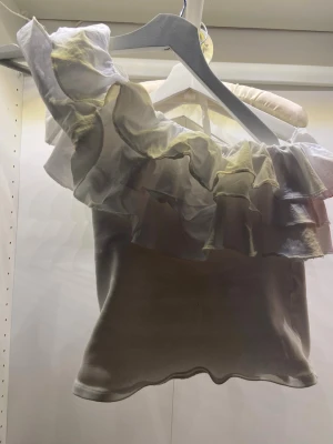 Vit oneshoulder topp med volanger Zara - Supersnygg vit oneshoulder topp från Zara med flera lager volanger över axeln. Tillverkad i ett mjukt material som känns bekvämt mot huden. Perfekt för dig som vill sticka ut med en unik och trendig topp. Använd två gånger