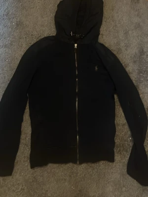 Blåa Ralph lauren hoodie med dragkedja - En svart hoodie med dragkedja framtill och huva. Klassisk design med långa ärmar och diskret logga på bröstet. Perfekt för en avslappnad stil och enkel att matcha med andra plagg.