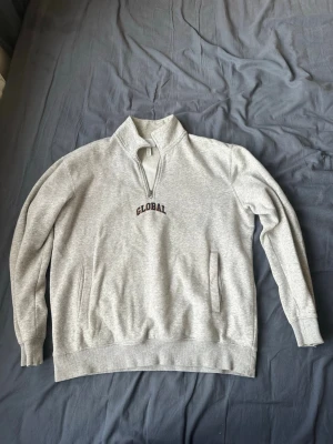 Grå halvzip sweatshirt med tryck - Ljusgrå sweatshirt med halv dragkedja och hög krage. På bröstet står det 'GLOBAL' i mörk text. Tröjan har två fickor framtill och ribbade muddar vid ärmar och nederkant. Tillverkad i mjukt sweatshirtmaterial, perfekt för en avslappnad stil.