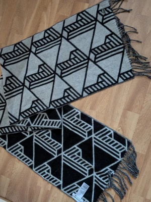 Mönstrad halsduk från Lacoste - Ladda upp inför vintern. Säljer en snygg halsduk från Lacoste med grafiskt svartvitt mönster och fransar i ändarna. Halsduken har en grå bas med svarta geometriska linjer och en liten Lacoste-logga. Perfekt accessoar för att lyfta din outfit.
