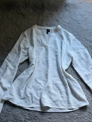 Vit långärmad blus från Vero Moda - Snygg vit blus från Vero Moda i storlek M. Blusen har lång ärm, v-ringad hals och är gjord i ett lätt, strukturerat tyg som ger en avslappnad men stilren look. Perfekt att matcha med jeans eller kjol för en trendig outfit.