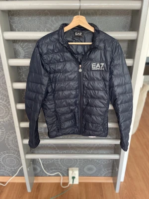 Svart dunjacka från Emporio Armani - Snygg svart dunjacka från Emporio Armani EA7 med quiltad design och dragkedja framtill. Jackan har två sidofickor med dragkedja och en hög krage. Perfekt för kalla vinterdagar och ger en stilren look.