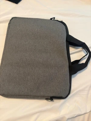 Grå laptopväska med handtag och axelband. - En stilren grå datorväska med svarta handtag och dragkedja. Väskan har en enkel, rektangulär form och är tillverkad i slitstarkt tygmaterial. Insidan är rymlig och fodrad, perfekt för att bära med sig laptop och tillbehör till skolan eller jobbet.