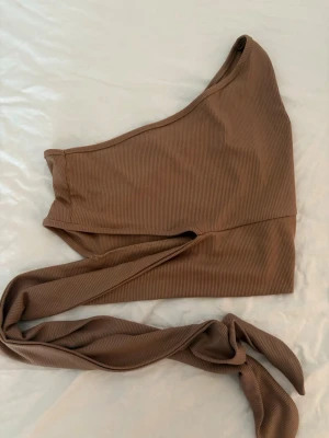 Brun oneshoulder topp med knyt - Säljer en brun ribbad oneshoulder topp med knyt i midjan. Toppen är ärmlös och har en snygg asymmetrisk skärning som ger en trendig look. Perfekt för dig som vill sticka ut lite extra med din outfit.