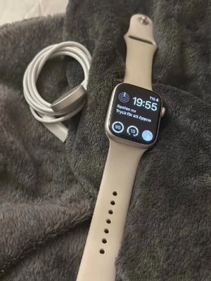 Apple Watch med beige sportarmband - Snygg Apple Watch med fyrkantig skärm och beige silikonarmband. Klockan har en modern design med touchskärm och kommer med laddare och originalförpackning. Perfekt för dig som vill ha koll på notiser, hälsa och träning direkt på handleden.