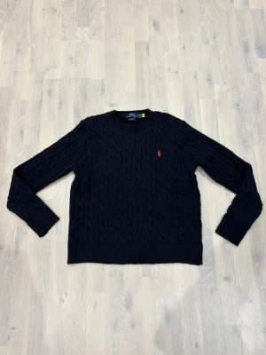 Mörkblå kabelstickad tröja Polo Ralph Lauren - Kabelstickad mörkblå tröja från Polo Ralph Lauren med klassisk rund halsringning och röd broderad logga på bröstet. Tröjan har långa ärmar och ribbade muddar vid ärmslut och nederkant. Perfekt för en stilren look.