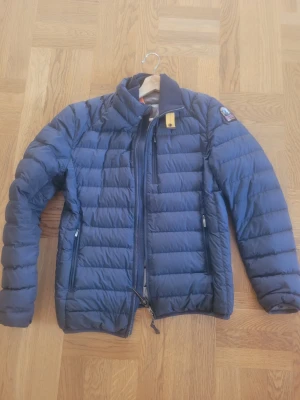 Blå parajumpers jacka - Snygg blå dunjacka med quiltad design och hög krage. Jackan har dragkedja framtill, två sidofickor med dragkedja och en patch på vänster ärm. Perfekt för kalla vinterdagar och riktigt lätt att bära.