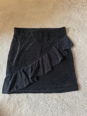 Svart glittrig kortkjol med volang - Snygg svart kortkjol med glittrigt tyg och en diagonal volangdetalj framtill. Kjolen har en tajt passform och passar perfekt när du vill sticka ut lite extra. Enkel att matcha med olika toppar för en trendig look.