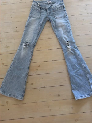 Ljusblå bootcut jeans STORLEK XS - Säljer ett par ljusblå bootcut jeans med slitningar på båda benen och cool tvätt. Jeansen har två stora framfickor och dubbla knappar i midjan. Perfekta för dig som gillar en avslappnad och trendig stil.