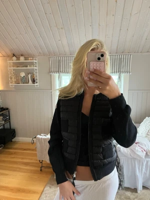 Svart pufferjacka med dragkedja - Snygg svart pufferjacka med quiltade partier och dragkedja framtill. Jackan har ribbade muddar vid ärmsluten och en kortare, croppad passform. Perfekt för att hålla värmen under kyliga dagar och ger en trendig look.