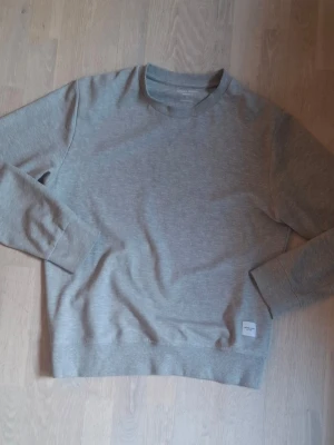 Ljusgrå basic sweatshirt - En stilren ljusgrå sweatshirt med rund hals och långa ärmar. Tröjan har ribbade muddar vid ärmslut och nederkant samt en liten vit patch nertill. Perfekt för en avslappnad look och enkel att matcha med andra plagg.