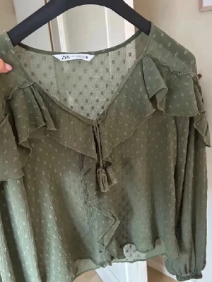 Olivgrön blus med volanger från Zara - Olivgrön blus från Zara med volangdetaljer längs ringningen och axlarna. Blusen har knytband med tofsar framtill och ett diskret prickmönster i tyget. Tillverkad i ett lätt, transparent material som ger en luftig känsla.