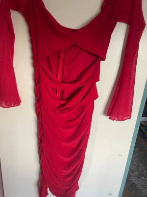 Röd långklänning med volang och mesh - Snygg röd långklänning med meshärmar och draperad kjol. Klänningen har volangdetaljer framtill och trekvartsärmar i transparent material. Perfekt för dig som vill sticka ut med en unik och trendig look.