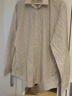 Blommig skjorta från Selected Homme - Säljer en vit skjorta från Selected Homme med små blommiga mönster i beige och blått. Skjortan har lång ärm, knappar framtill och en slim fit passform. Tillverkad i ett lätt och bekvämt bomullsmaterial som är enkel att stryka.