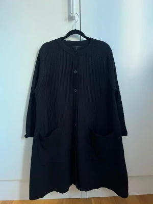 Svart lång kofta från COS - Säljer en svart lång kofta från COS med rund halsringning och knäppning framtill. Koftan har två stora fickor fram och är tillverkad i ett strukturerat, stickat material. Perfekt för lager på lager och en clean look.