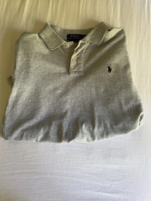 Grå pikétröja från Polo Ralph Lauren - Klassisk grå pikétröja från Polo Ralph Lauren med korta ärmar och krage. Tröjan har knappar vid halsen och den ikoniska broderade loggan på bröstet. Perfekt till jeans eller shorts för en clean och stilren look.