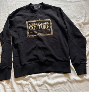 Svart sweatshirt Versace Jeans Couture - Svart sweatshirt från Versace Jeans Couture med guldfärgat tryck framtill. Klassisk rund halsringning och ribbade muddar vid ärmslut och nederkant. Perfekt för en trendig och avslappnad look.