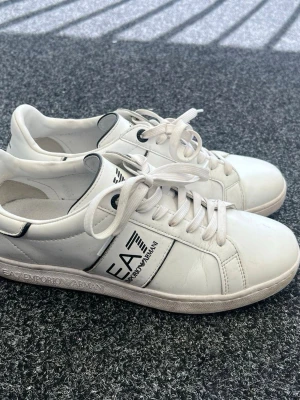 Vita EA7 Emporio Armani sneakers - Snygga vita sneakers från EA7 Emporio Armani med svart logga på sidan och detaljer i svart. Skorna har klassisk låg profil, vita snören och är tillverkade i slätt lädermaterial. Perfekta för en clean och stilren look. Pris kan förhandlas.