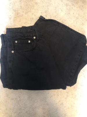 Svarta baggy jeans med slitna byxben - Säljer ett par svarta jeans med baggy passform och rå, fransad kant längst ner på benen som blivit pågrund av välanvändning. Jeansen har klassisk femficksdesign och silverfärgade nitar. Materialet är denim i bomull. Perfekta för en avslappnad och trendig look. I ganska slitet skick men har fortfarande mer att ge, därav det billiga priset.