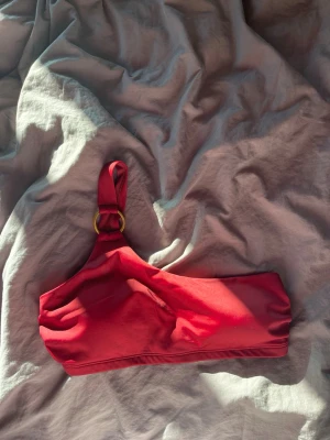 Röd bikiniöverdel med ringdetalj - Snygg röd bikiniöverdel med en axel och guldfärgad ringdetalj på axelbandet. Trendig asymmetrisk design som ger en cool look på stranden eller vid poolen. Tillverkad i mjukt och stretchigt material för skön passform.