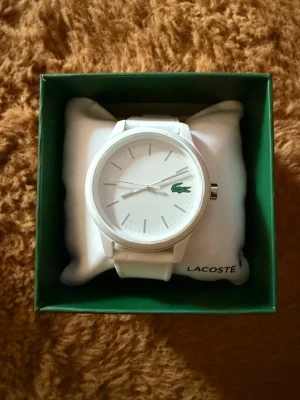 Vit klocka från Lacoste - Stilren vit klocka från Lacoste med minimalistisk design och klassisk rund urtavla. Klockan har ett vitt armband och den ikoniska gröna krokodilen på urtavlan. Perfekt accessoar för dig som gillar enkel och modern stil.