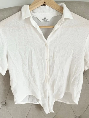 Vit kortärmad skjorta med knyt - Säljer en vit kortärmad skjorta från H&M i ett lätt och luftigt viscose tyg. Skjortan har klassisk krage, knappar framtill och en snygg knytning nedtill för en avslappnad look. Perfekt till sommaren och enkel att styla till olika outfits.